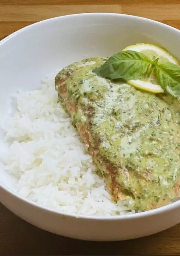 Salmon in creamy pesto sauce on white rice, garnished with lemon and basil in a white bowl on a wooden table.